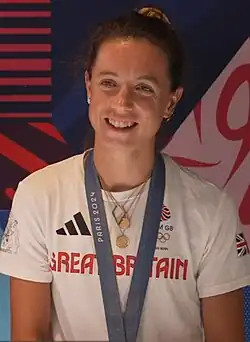 Imogen Grant trägt ein weißes Hemd mit der Aufschrift Great Britain. Um den Hals trägt sie einige Ketten und ein blaues Band, an dem ihre Goldmedaille befestigt ist, die aber nicht im Bild ist.