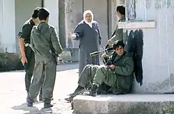 eine alte Frau mit Kopftuch spricht mit Jugendlichen, die Gewehre in den Händen halten