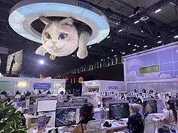 Das Bild zeigt den Stand der Entwicklerfirma auf der Gamescom 2024. In der unteren Bildhälfte probieren Besucher das Spiel an Computern aus, in der oberen Bildhälfte schwebt eine große Lampe in Ringform (wie das o aus dem inZOI-Logo) in der die überdimensionale Plüschkatze Psycat hängt.