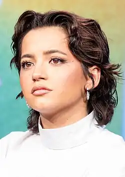 Isabela Merced auf der SXSW 2025. Sie hat mittellanges, welliges, dunkelbraunes Haar, das locker gestylt ist. Sie trägt einen weißen Rollkragenpullover, der elegant und schlicht wirkt, sowie silberne Ohrringe. Der Hintergrund ist unscharf und zeigt eine grüne und gelbe Farbgebung.