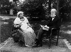 Das Schwarz-Weiß-Bild zeigt das Ehepaar Hooker in einem Garten. Während im Hintergrund links eine architektonische Struktur und rechts Bäume zu sehen sind, sitzt das Ehepaar im Vordergrund auf zwei Holzstühlen, die auf einen auf dem Rasen ausgelegten Teppich stehen. Beide sind vollständig zu sehen. Beecher Hooker sitzt links, blickt nach rechts, hat weißes, krauses Haar trägt ein helles Kleid mit Spiralenmuster, einen weißen Umhang und sitzt auf einer dunklen Decke, auf der sich teilweise andersfarbige Streifen abzeichnen. Soweit erkennbar, hält sie in ihrer rechten Hand ein Stück Papier. Rechts von ihr sitzt ihr Ehemann, der nach hinten gekämmtes graues Haar und einen halbwegs langen grauen Vollbart hat. Er blickt nach links, trägt ein weißes Hemd mit dunkler Fliege, Weste und Anzug, grauer Hose und schwarzen Schuhen. Mit seinen Händen hält er auf seinem Schoß einen aufgeschlagene Zeitung oder ein aufgeschlagenes Buch fest.