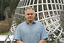 Dieses Farbfoto zeigt József Balogh vor einen Kunstobjekt stehend als Frontalaufnahme einschließlich Brustkorb im Forschungszentrum Oberwolfach für "Kombinatorik" im Jahr 2023.