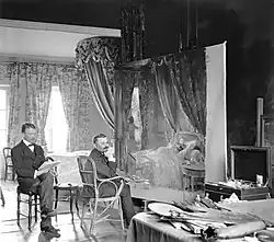 Muenier beim Malen des Bildes „Das Erwachen“ bei seinem Sohn Pierre. Die beiden Männer sitzen vor einer großen Leinwand in einem Schlafzimmer mit hohen Decken und einem luxuriösen Himmelbett.