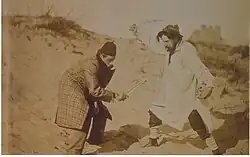 Sepia-Fotografie von zwei kostümierten Männern in einer Dünenlandschaft, die miteinander zu fechten scheinen. Der linke trägt einen karierten Mantel und einen Fez, der rechte einen hellen Kittel und eine Mütze. In den Händen halten sie Knochen und einen Totenschädel.