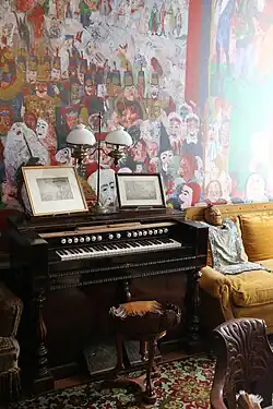 Farbfotografie des restaurierten James-Ensor-Hauses. Hinter einem Harmonium und einem Sofa ist an der Wand ein Ausschnitt des Gemäldes „Der Einzug Christi in Brüssel“ zu sehen.