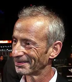 James Kent steht auf einem roten Teppich. Im Hintergrund ist ein Absperrzaun zu sehen, an dem einige Personen stehen. Kent trägt ein schwarzes Jackett, ein weißes Hemd, seine dünnen, grauen Haare bis zu den oberen Ohren sowie einen stoppeligen Kinn- und einen dünnen Schnurrbart.