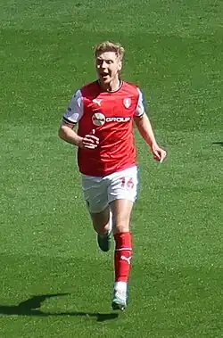 Fußballer im Sportkleidung von vorne in Laufbewegung auf grünen Rasen.