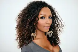 Porträtfoto von Janet Mock vor weißem Hintergrund. Sie trägt ein graues Oberteil, Wimperntusche, goldfarbene, große Ohrringe mit in der Mitte runterhängenden Spitzen und ihre braunen Haare als Locken.