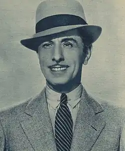 Mann ab Brustkorb von vorn im hellen Anzug und Hemd nebst gestreifter Krawatte und hellem Hut nebst schwarzem Streifen über der Hutkrempe in schwarzweiß.