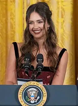 Jessica Alba spricht im Weißen Haus, 18. September 2024. Sie steht hinter einem Podium mit dem Siegel des Präsidenten der Vereinigten Staaten. Der Hintergrund besteht aus einem gelben Vorhang, der eine helle und formale Atmosphäre erzeugt. Jessica Alba hat langes, welliges Haar und trägt ein ärmelloses Kleid mit einem Blumenmuster. Auf dem Podium sind zwei Mikrofone angebracht.