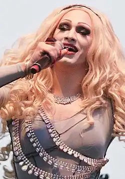 Jinkx Monsoon singt in ein Mikrofon, das sie in der rechten Hand hält. Am Mittelfinger derselben Hand trägt sie einen orangenen Ring mit rotem Rubin in der Mitte, am Arm zudem einige Reife. Sie trägt eine blonde Perücke, dunklen Lidschatten, falsche Wimpern, aufgemalte Augenbrauen, roten Lippenstift, rot lackierte Fingernägel, eine Diamant-Halskette und ein beiges Kleid, über das Edelstein-Ketten wie Bänder oder Gürtel gespannt sind.
