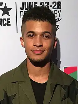 Jordan Fisher steht vor einer weißen Wand mit der schwarzen Aufschrift June 23-26. Er trägt eine dunkelgrüne Jacke mit schwarzen Sternen, ein schwarzes T-Shirt und einen leichten Stoppelbart am Kinn. Sein schwarzen Haare sind an den Seiten bis zur Stirn rasiert, darüber leicht gekräuselt frisiert.