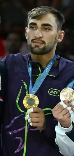 Kotsoiev trägt einen dunkelblauen Trainingsanzug, die Goldmedaille ist an einem hellblauen Band um seinen Hals befestigt.