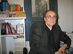 Ein Mann Anfang 60 mit heller Haut, grauen Haaren und einer Halbglatze mit Brille. Er trägt einen dunkelgrauen Anzug und ein schwarzes Hemd. Im Hintergrund ein weißes Regal mit Fachbüchern und ein blaues Fenster.