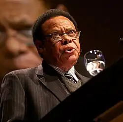 Juma Mwapachu. 2012. Ein afrikanischer Mann mit kurzem Kraushaar und Brille hinter einem Wasserglas an einem Rednerpult mit einer Großprojektion auf der Leinwand hinter ihm.