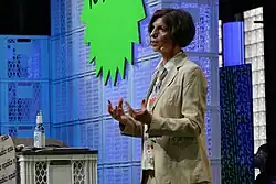 Jutta Allmendinger spricht auf der re:publica 2023, die an einem Podium steht. Sie trägt eine beige Jacke über einem bunten Hemd und hat kurze Haare. Das Podium besteht aus weißen Plastikkisten, auf denen eine Wasserflasche steht. Hinter ihr befindet sich ein großes grünes Schild mit gezackten Kanten, auf dem „re:publica“ zu lesen ist. Der Hintergrund zeigt gestapelte Plastikkisten und ein blaues Schild mit dem wiederholten Schriftzug „CASH“ in blauen Buchstaben.