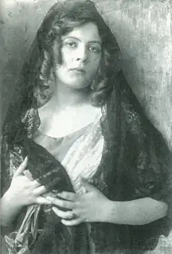 Eine junge Frau mit langen dunklen Haaren, über die ein langer schwarzer Schleier gelegt ist, den sie in Brusthöhe mit einer Hand umfasst