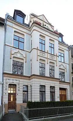 Mehrstöckiges Wohngebäude mit einer kunstvollen Architektur. Die Fassade des Gebäudes ist in Beige- und Weißtönen gehalten und weist dekorative Elemente wie aufwendige Verzierungen und Muster um die Fenster und Türen auf. Die Fenster sind groß und mit dunklen Holzrahmen versehen. Vor dem Gebäude befindet sich ein kleiner Garten mit Hecken, der von einem Metallzaun umgeben ist. Der Eingang ist aus Holz und hat Glaspaneele, mit einer kleinen Treppe, die hinaufführt. Das Gebäude wirkt gepflegt und strahlt einen klassischen, eleganten Stil aus.