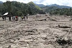 Eine von einer Schlammlawine gefüllte Fläche auf der links eine Gruppe Männer und Kinder steht. Am Rand sind zerstörte Hütten zu erkennen. Im Hintergrund befinden sich mit Gras und Bäumen bewachsene Berge.