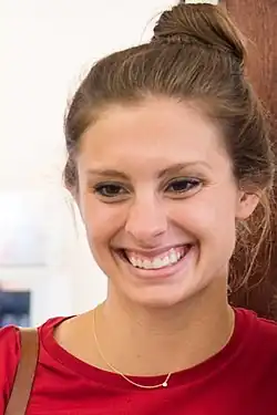 Die lächelnde Katie Meili trägt ihr Haar hier hochgesteckt, sie hat ein rotes Hemd an. Auf der rechten Schulter ist ein braunes Lederband erkennbar, vermutlich der Trägergurt einer Tasche.