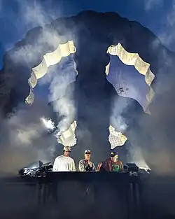 DJs Adam Port, &ME und Rampa auf einer Bühne an einem DJ Pult. Im Hintergrund ein großes Peace Symbol.