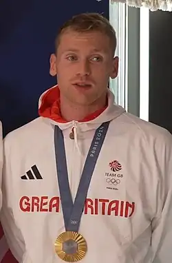 Bird trägt während eines Interviews eine weiße Trainingsjacke. An einem blauen Band um seinen Hals ist seine olympische Goldmedaille befestigt.