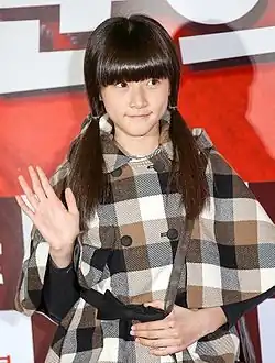 Kim Sae-ron mit langen, glatten Haaren, die zu zwei Zöpfen gestylt sind. Sie trägt einen karierten Mantel mit einem Muster aus großen Quadraten in Braun-, Schwarz- und Weißtönen. Sie winkt mit der linken Hand, während die rechte Hand auf ihrem Bauch ruht. Der Hintergrund ist rot mit einigen weißen und dunklen Elementen, die möglicherweise zu einem Schild oder Poster gehören.