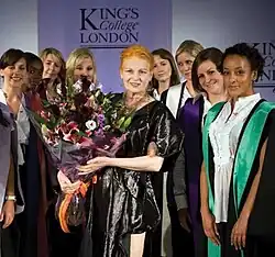Frontale Farbfotografie von Westwood mit einem großen Blumenstrauß an ihrer rechten Seite. Sie ist von einer Gruppe junger Frauen umgeben, die verschiedenfarbige Talare tragen. Im Hintergrund ist die Aufschrift King’s College London zu erkennen.