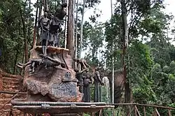 Das Foto im Querformat zeigt ein großes Denkmal. Rechts im Hintergrund ist ein Wasserfall zu erkennen, der eine braune Felsklippe hinunterfällt. Rundherum stehen Bäume und links hinter dem Denkmal führt ein Fußweg eine Treppe hinauf. Das Denkmal selbst zeigt drei Menschen auf einer Klippe. Einer dieser ist ein Mann mit grimmigem Gesichtsausdruck und einem Speer in der rechten Hand. Seine linke Hand ist abweisend nach vorne gerichtet. Vor ihm steht an der Klippenkante eine gefesselte Frau, deren Rücken bereits über diese lehnt, da sie vermutlich gerade über diese geschubst wurde. An der Klippe hängend befindet sich eine weitere gefesselte Frau mit leidvollem Gesichtsausdruck und weit aufgerissenem Mund. In die Klippe eingraviert ist zudem auf der rechten Seite ein Wasserfall und darunter ein Kreuz mit aufgeschlagenem Buch und eine Frontalansicht des Kisiizi-Krankenhauses. Rechts neben der Felsenklippe stehen als Teil des Denkmals drei Frauenfiguren, die lächelnd auf ein Kind blicken, das die links stehende Frau in die Höhe gehoben hat. Eine der anderen beiden Frauen scheint schwanger und die dritte eine Krankenschwester zu sein.