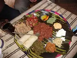 Auf einem geflochtenen Teller auf einem grob gewebtem, gestreiftem Tischtuch. Auf dem Teller liegen auf großen Blätter Rinderhack und darum herum Teigröllchen, geröstetes Fladenbrot und Häufchen Pasten in verschiedenen Tönungen von weiß, braun und grün und ein Schälchen mit Nitir qibe