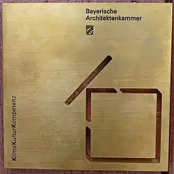 Messingfarbenes Quadrat mit einem stilisierten Haus in der rechten unteren Ecke. Oben rechts steht Bayerische Architektenkammer, darunter ist das bayerische Wappen. Am unteren linken Rand geht der Schriftzug Klimakulturkompetenz nach oben.