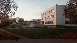 Rechte Bildhälfte: Quaderförmiger mit Erdgeschoss, zwei Obergeschossen und einem Untergeschoss. Links: ansteigender, kurviger Weg zu einem eingeschossigen Eingangsbereich mit großen Fensterflächen, das am größeren Gebäudeteil angeschlossen ist. Vor dem Eingangsbereich steht ein Auto. Menschen sind nicht zu sehen.