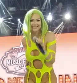 Porträtfoto von Kylie Sonique Love, die ein schwarzes Mikrofon in der linken Hand hält. Sie trägt ein über den Oberkörper gespanntes, dünnes, löchriges, gelbes Gummiteil und eine hellgrüne, runde Perücke, die bis zu den Schultern reicht. Im Hintergrund ist eine hellrötlich-weiße Wand mit der weißen Aufschrift Cowboy Music Festival Calgary zu sehen. Die Aufschrift ist in einem aufgemalten Saloon-Schild mit einem oberen Stern angebracht. Darunter sind verdeckte rote Neon-Buchstaben zu sehen. An der Decke hängen einige schwarze Scheinwerfer.