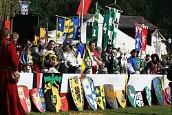 Das Foto zeigt viele bunte, an einem langen Tisch aufgereihte Wappenschilde und dahinter Personen in Ritterrüstungen, die ihre verzierten Helme vor sich abgelegt haben.