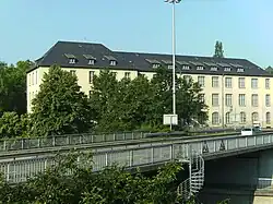 Eines der ursprünglichen Magazingebäude. Ansicht vom Nordostrand des Langemarckplatzes außerhalb der Liegenschaft aus, Blickrichtung: südwest, die auf dem Bild sichtbare lange Seite des Gebäudes zeigt Richtung Nordosten; die über die Brücke im Vordergrund führende Straße ist die B 416 Mayener Straße, das silberne Auto fährt Richtung Westen, unter der Brücke hindurch führt die B 9. (Die unten dargestellte Photopostkarte zeigt dieses Gebäude vor Kriegsbeschädigung und Wiederaufbau).