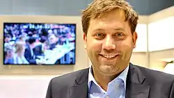 Ein lächelnder Mann im Anzug steht in einem gut beleuchteten Raum. Ein verschwommener Bildschirm hinter ihm zeigt eine Menschenmenge bei einer Konferenz. Die Atmosphäre ist professionell und fröhlich.