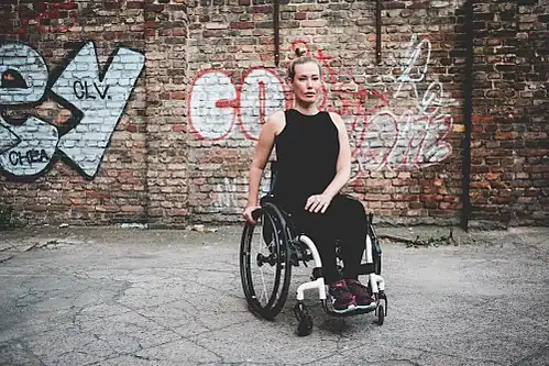 Laura Gehlhaar ist vor einer Ziegelmauer, die mit Graffiti besprüht ist abgebildet. Ihr Haar ist blond und zurückgebunden, sie trägt dunkle Kleidung und ihr Rollstuhl ist schwarz.