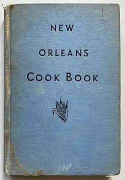 Ein hellblaues Buch liegt auf einem weißen Untergrund. In der Buchmitte steht in schwarzen Großbuchstaben über drei Zeilen verteilt der Titel New Orleans Cook Book. Etwas unter dem Schriftzug Cook Book befindet sich eine kleine, ebenfalls schwarze Zeichnung von zwei Maiskolben. Die Buchränder sowie der Buchrücken sind teils deutlich abgenutzt.