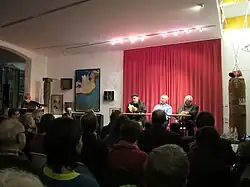 Drei Autoren auf einer Bühne vor einem roten Vorhang sitzend, im vorderen Bildbereich Publikum von hinten