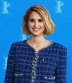 Linda Caridi auf der Berlinade 2023, trägt eine Jacke mit einem karierten Muster in verschiedenen Blautönen, die durch einen dekorativen Rand entlang des Kragens und der Knopfleiste hervorgehoben wird. Dieser Rand ist mit Perlen und anderen Verzierungen gestaltet. Auf der linken Seite der Jacke ist ein gelbes Abzeichen zu sehen, das einen markanten Kontrast zum Blau bildet. Der Hintergrund des Bildes ist in einem tiefen Blau gehalten und zeigt verschwommene weiße Silhouetten vom Berliner Bären der Berlinade. Die gesamte Komposition erzeugt eine Mischung aus Struktur und Eleganz, wobei die dekorativen Details der Jacke besonders ins Auge fallen.