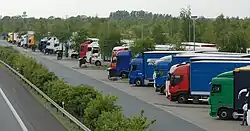 Mehrere Lkws stehen nebeneinander auf einer Betonfläche, dazwischen und dahinter stehen Bäume. Durch eine Leitplanke und eine Reihe Büsche ist die Fläche mit den Lkws von der Autobahn abgetrennt.