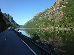 Bild von einer Straße, die entlang eines Gewässers führt. Links und rechts erheben sich teils begrünte, teils felsige Berge.