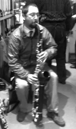 Sitzender Mann in legerer Kleidung mit hellem Hemd, dunkler Krawatte, heller Hose und Freizeitjacke sowie einem länglichen flötenartigen, länglichen und verschnörkelten Instrument zwischen den Beinen, welches er bläst in schwarzweiß.