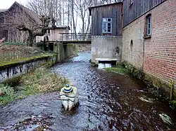 Ehemalige Wassermühle an der Lutter in Marwede, mit Steinskulptur “Die Heide aus Lüneburg” von Petra Förster