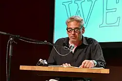 Oberkörper eines sitzenden Mannes mit kurzen grauen Haaren und dickrandiger Brille, der in ein Standmikrofon spricht. Vor ihm ein Holztisch mit einem aufgeschlagenen Buch in das er blickt.