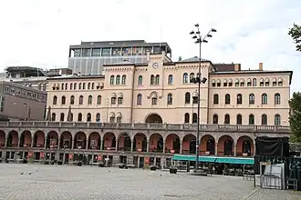 Foto eines Gebäudes mit verzierter Fassade und einer Uhr darin, davor ein Platz