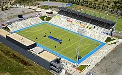 Ein Hockeystadion aus der Vogelperspektive. Umgeben ist das grüne Spielfeld von einem blauen Streifen, außen herum stehen Zuschauertribünen