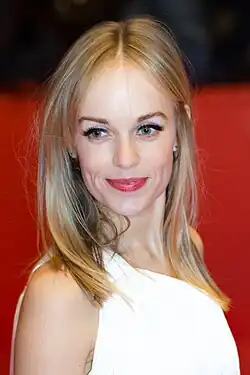 Friederike Kempter mit schulterlangem, blondem Haar und ein weißes ärmelloses Oberteil trägt. Der Hintergrund ist ein einheitliches Rot, Sie lächelt in die Kamera und hat einen natürlichen Make-up-Look. Das Bild entstand auf dem rotem Teppich der Berlinale 2017.