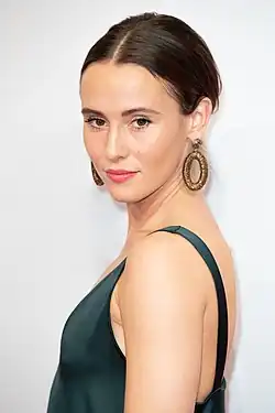 Peri Baumeister in einem eleganten, dunkelgrünen, ärmellosen Kleid. Ihr braunes Haar ist mittig gescheitelt und nach hinten gestylt, auf der Medienboard Berlin-Brandenburg Party zur 69. Berlinale. Sie trägt auffällige, ovale, goldene Ohrringe. Der Hintergrund ist schlicht und weiß.