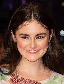 Lea van Acken lächelt auf der Berlinale 2020 mit langem, dunkelbraunem Haar, das in der Mitte gescheitelt ist. Sie trägt auffälligen Ohrschmuck an beiden Ohren, darunter kleine Stecker und Kreolen. Sie ist in ein farbenfrohes Oberteil gekleidet, das ein florales Muster und funkelnde Pailletten aufweist. Der Hintergrund des Bildes ist dunkel, was darauf hinweist, dass es entweder nachts oder in einer schwach beleuchteten Umgebung aufgenommen wurde.